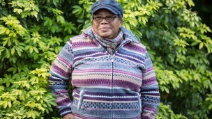 A woman in glasses and a hat looks directly at the camera with a tree in the background